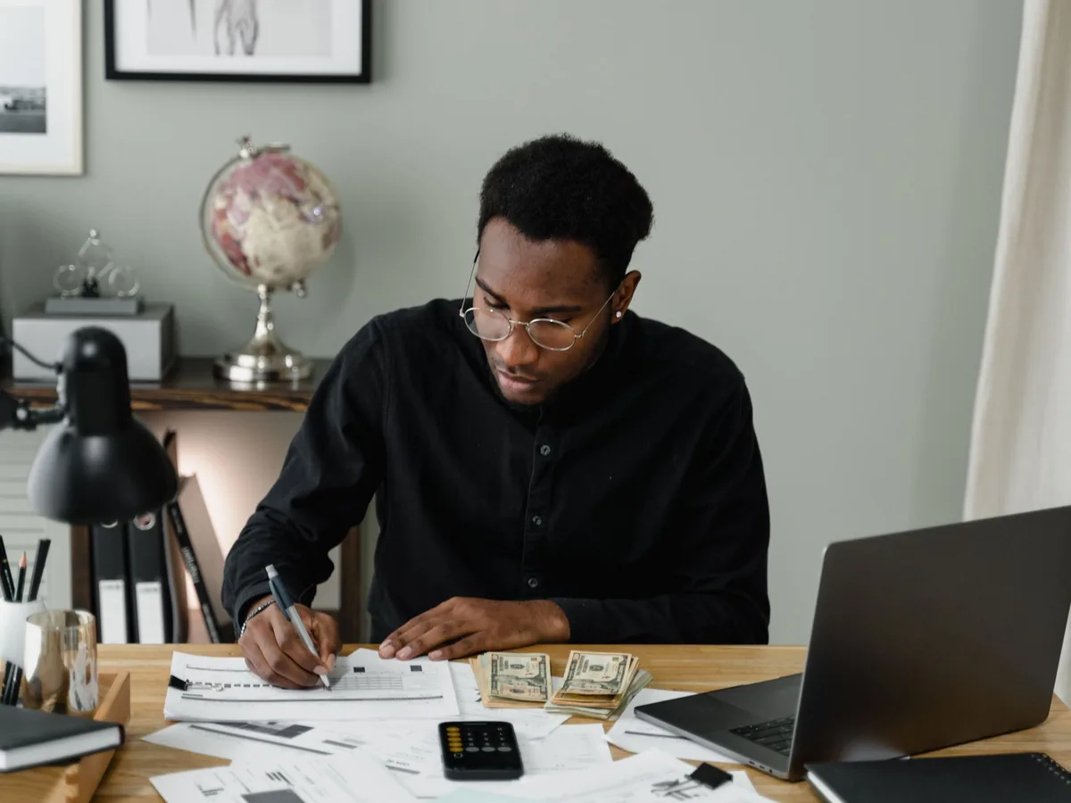 A man writing on documents at a desk with stacks of banknotes, a calculator and a laptop