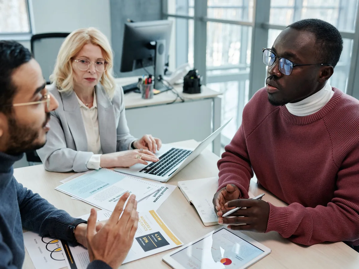 Three colleagues talking at a meeting table with a laptop, a tablet and printed reports