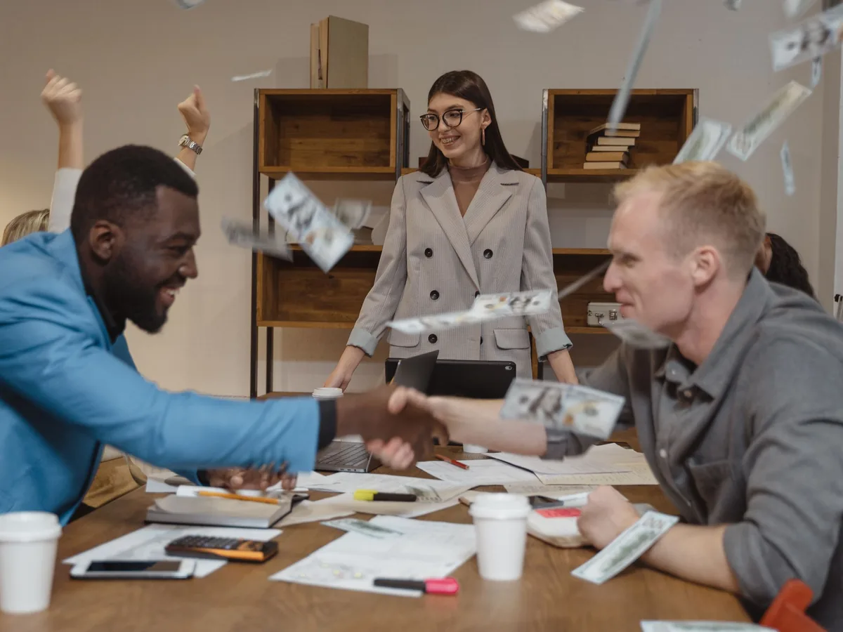 Colleagues shaking hands across a meeting table as banknotes fall through the air