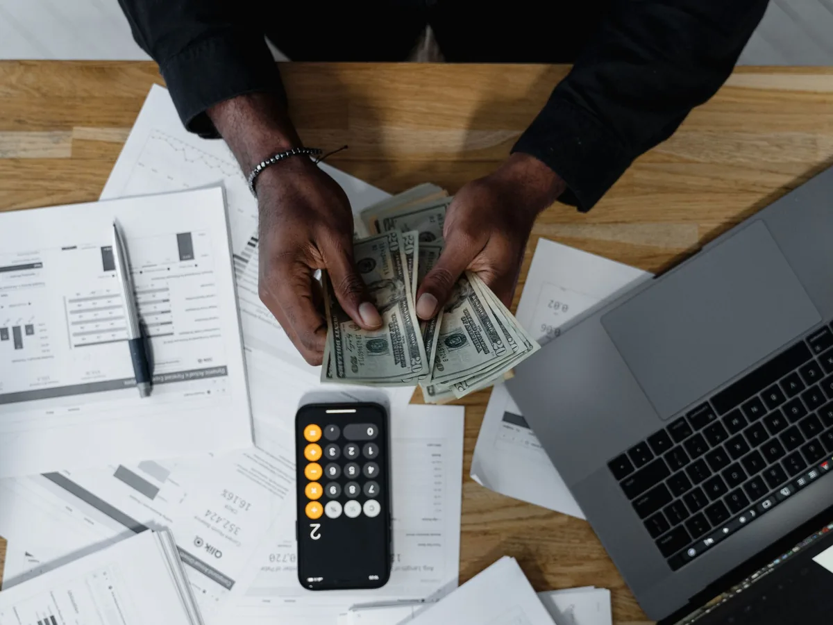 Overhead view of hands counting banknotes beside a laptop, a phone calculator and printed charts